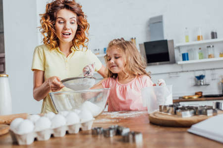 selective focus of curly woman sieving flour into glass bowl near daughter, chicken eggs and cookie cuttersの写真素材
