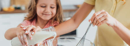 cropped view of mother holding whisk and pouring milk into glass bowl together with daughter, panoramic shotの写真素材