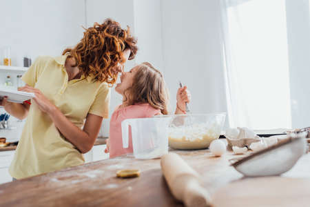 selective focus of mother holding plates while standing face to face with daughter near table with cooking utensilsの写真素材
