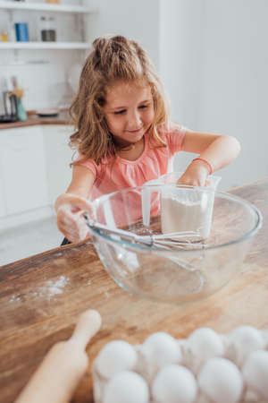 selective focus of child mixing flour in measuring jug near glass bowl, rolling pin and chicken eggsの写真素材