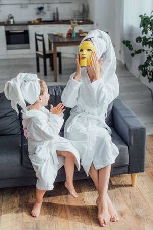 high angle view of woman putting on face mask while sitting with daughter in bathrobes and towels on headsの写真素材