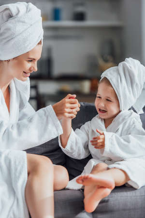 selective focus of young mother making manicure to child while sitting together in white bathrobes and towels on headsの写真素材