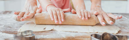 partial view of woman and child rolling out dough near cookie cutters on kitchen table, panoramic...