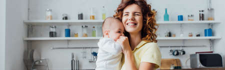 panoramic shot of young mother with closed eyes holding infant boy in kitchenの写真素材