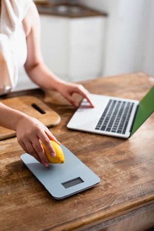cropped view of woman holding lemon near kitchen scales and using laptop