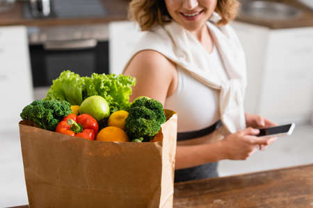 selective focus of paper bag with groceries near woman holding smartphone with blank screenの写真素材