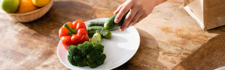 panoramic orientation of young woman holding cucumber near plate with broccoli and bell pepper