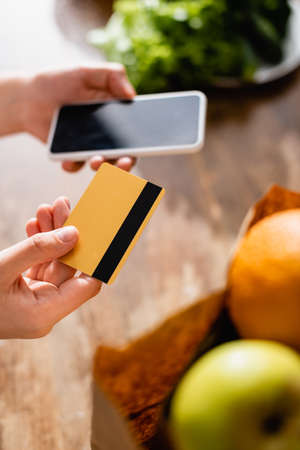 selective focus of woman holding credit card and smartphone with blank screen near fruits in paper bagの写真素材