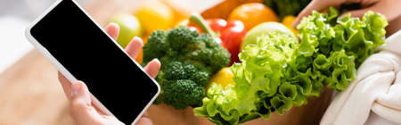 panoramic crop of woman holding smartphone with blank screen near groceriesの写真素材