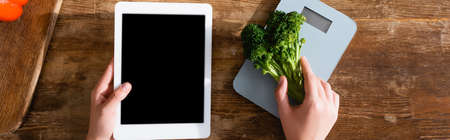 horizontal image of woman holding broccoli near kitchen scales and digital tablet with black screenの写真素材