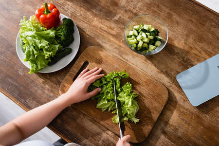 top view of woman cutting green lettuce on chopping board near kitchen scales and whole vegetablesの写真素材