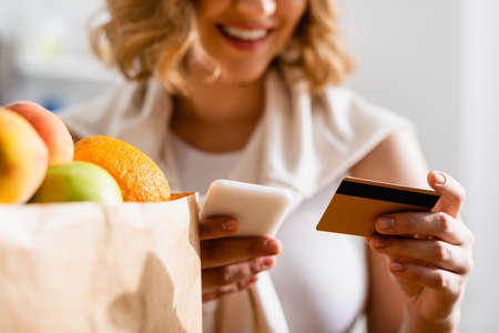 partial view of woman holding smartphone and credit card near fruits in paper bag