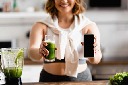 cropped view of young woman holding glass of green smoothie and smartphone with blank screenの写真素材