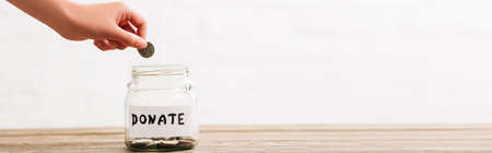 cropped view of woman putting coin in penny jar with donate lettering on wooden surface on white background, panoramic shotの写真素材
