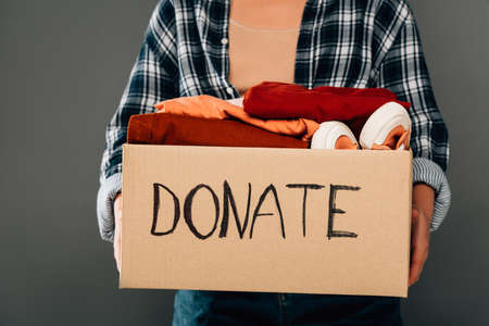 cropped view of woman holding box with donate lettering and clothes on gray backgroundの写真素材