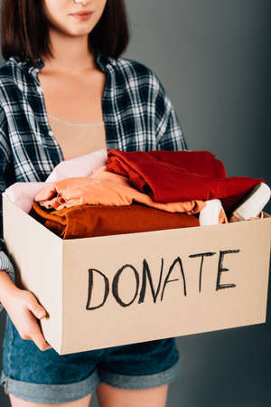 cropped view of woman holding box with donate lettering and clothes on gray backgroundの写真素材