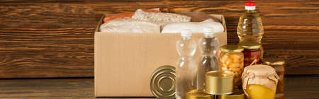 panoramic shot of cardboard box with groats near water, oil, canned food and honey on wooden...