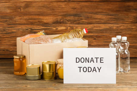 cardboard box with groats near water, oil, canned food, honey and donate today card on wooden background, charity conceptの写真素材