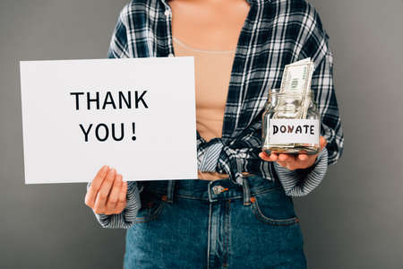 cropped view of woman holding card with thank you lettering and jar with donated money on gray backgroundの写真素材