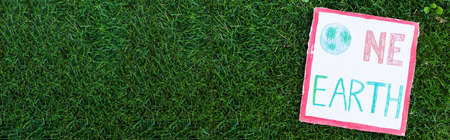 Panoramic shot of placard with one earth lettering on green grass outdoors, ecology concept