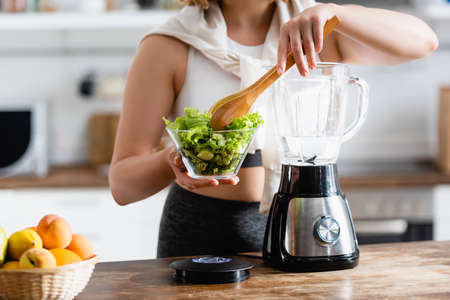 cropped view of woman holding bowl with lettuce and wooden spoon near blender