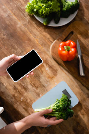 top view of woman holding broccoli near kitchen scales and smartphone with blank screenの写真素材