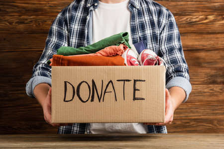 cropped view of man holding cardboard box with donate lettering and clothes on wooden background, charity conceptの写真素材