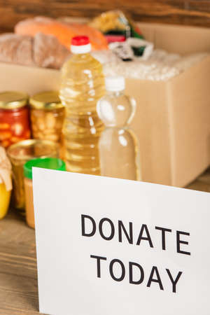 selective focus of cardboard box with food and donate today card on wooden background, charity conceptの写真素材