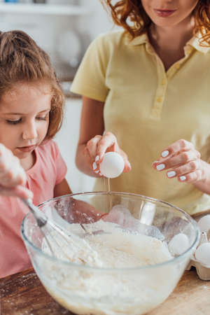 partial view of mother holding chicken egg near daughter kneading dough in glass bowl