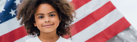 panoramic crop of curly african american kid looking at camera near flag of america