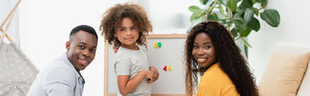 panoramic crop of african american family looking at camera near magnetic easelの写真素材