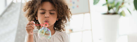 panoramic shot of curly african american kid blowing soap bubbles