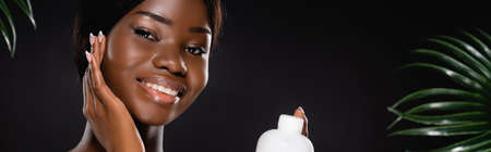 african american woman holding bottle of lotion near green palm leaves isolated on black, panoramic...