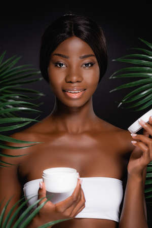 african american woman with cosmetic cream near green palm leaves isolated on blackの写真素材