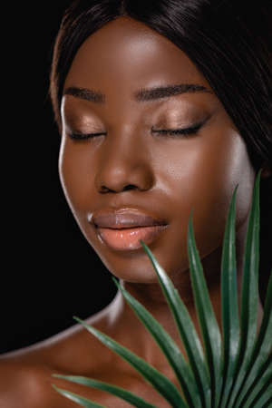 african american naked woman with closed eyes and green palm leaf isolated on blackの写真素材