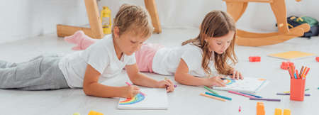 concentrated brother and sister in pajamas lying on floor and drawing with felt pens in sketchbooksの写真素材