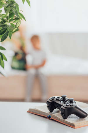 KYIV, UKRAINE - JULY 21, 2020: selective focus of joystick on open book, and boy sitting on bed near...