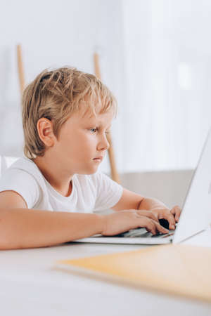 selective focus of focused kid in white t-shirt using laptop while sitting at table