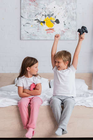 boy showing winner gesture while holding joystick near offended sister sitting with crossed armsの写真素材