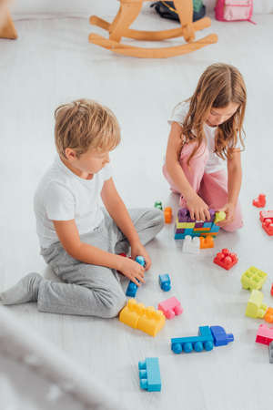 high angle view of concentrated siblings in pajamas sitting on floor and playing with building blocksの写真素材