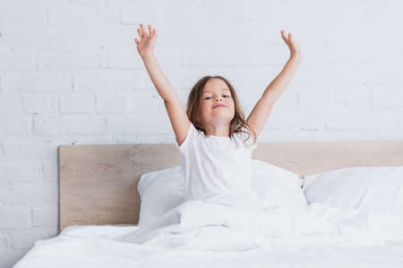 awakened girl stretching while sitting in bed and looking at cameraの写真素材