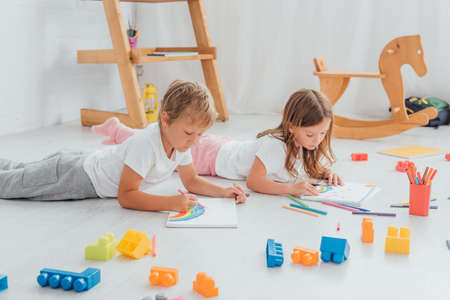 brother and sister in pajamas lying on floor near building blocks and drawing with felt pens