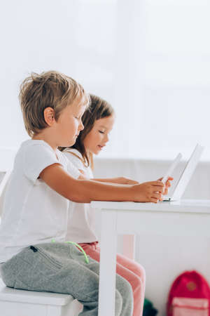 side view of brother and sister in pajamas sitting at while table and using laptops togetherの写真素材