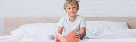 horizontal concept of boy in white t-shirt looking at camera while sitting in bed with book