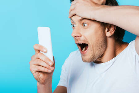 Shocked man with hand near head looking at smartphone on blue background