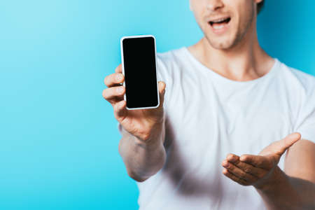 Cropped view of man pointing with hand while showing smartphone with blank screen on blue background