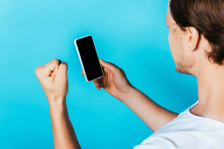 Man showing yeah gesture and holding smartphone with blank screen on blue backgroundの写真素材