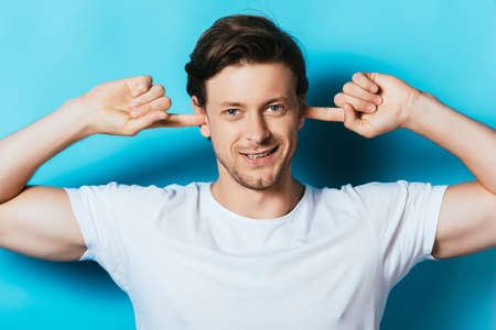 Young man in white t-shirt covering ears with fingers on blue background