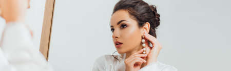Panoramic shot of bride in silk robe putting on pearl earring near mirror