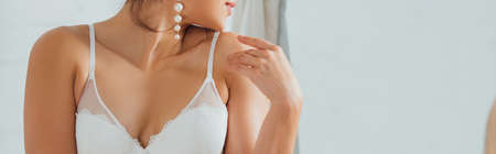 Panoramic shot of sexy bride in white bra and pearl earring touching shoulder at homeの写真素材